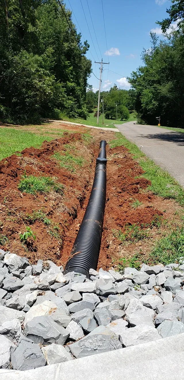 Black drainage pipe in a trench beside a gravel-lined road. Lush green trees frame a blue sky.