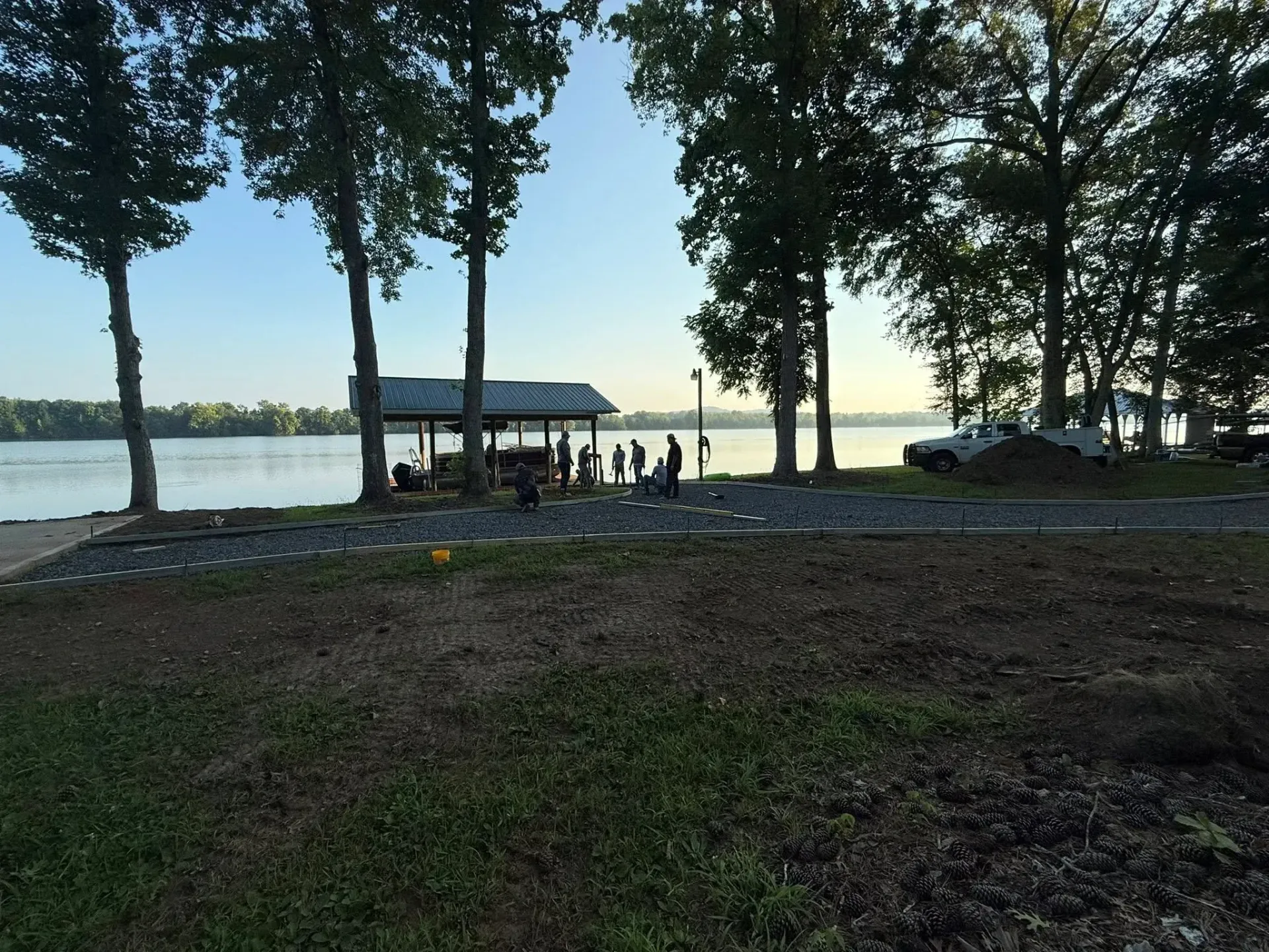 People gathered near a covered structure beside a lake, trees line the shore, daytime.