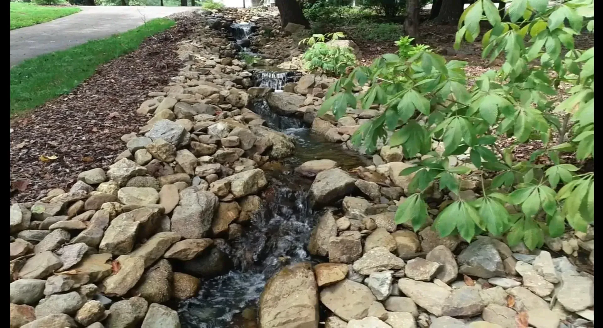 A stream lined with rocks flows through a garden, with green plants and a walking path visible.