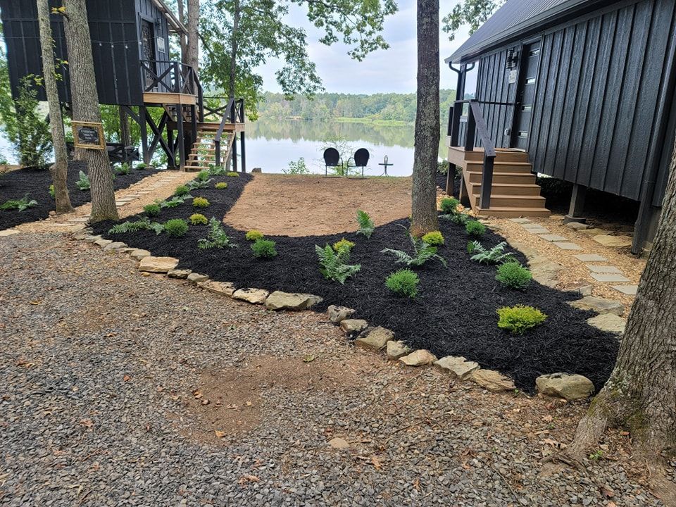 Black cabins by a lake, surrounded by landscaping with gravel driveway and mulched garden beds.