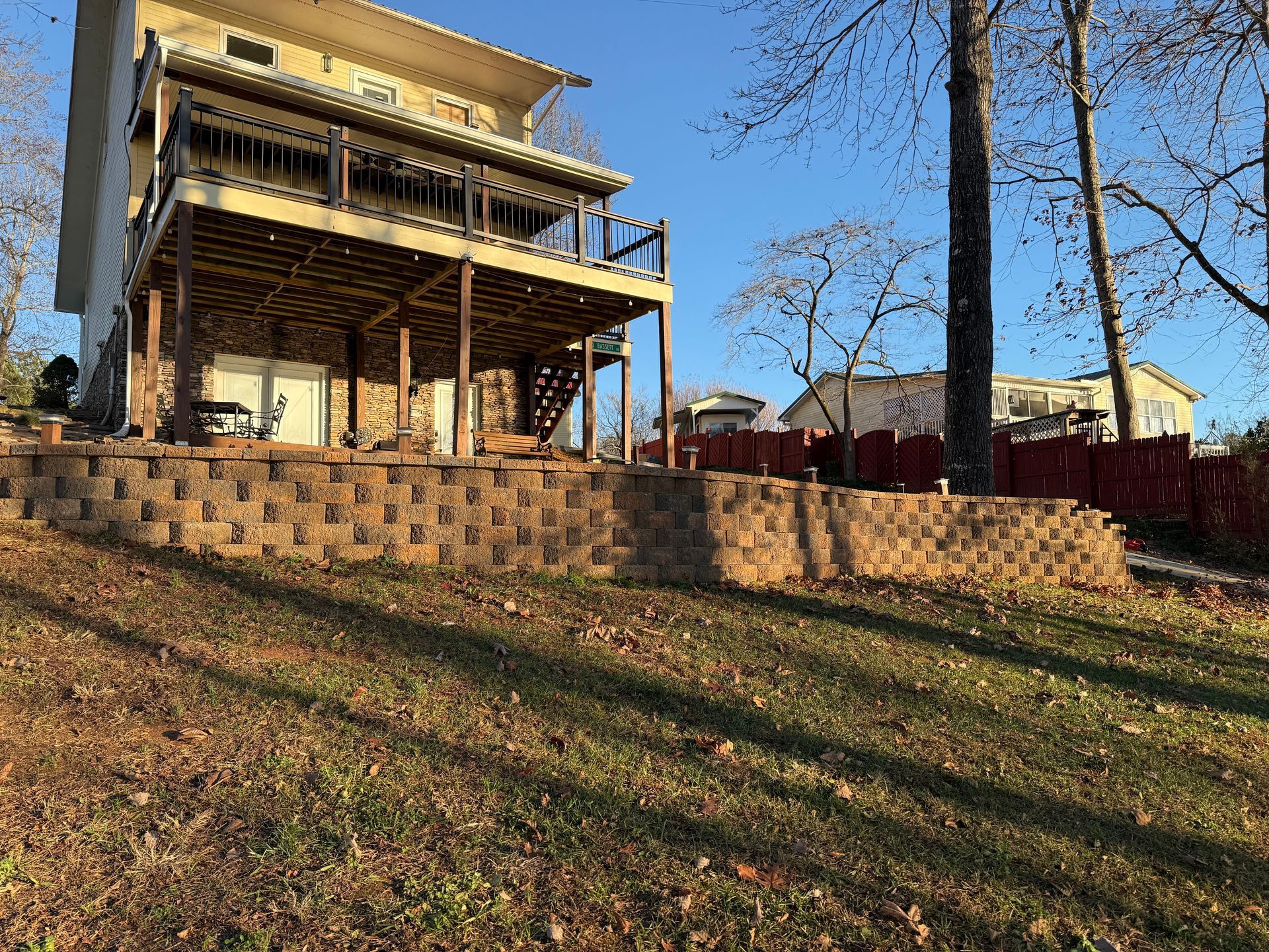 A two-story house with a wooden deck, built on a block retaining wall on a grassy slope.