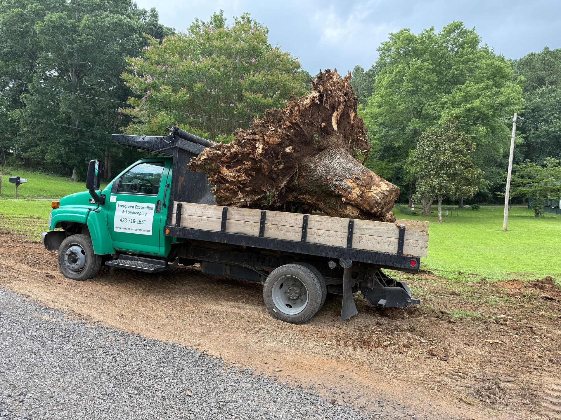 Green truck carrying a large tree stump on a gravel road next to a green field and trees.