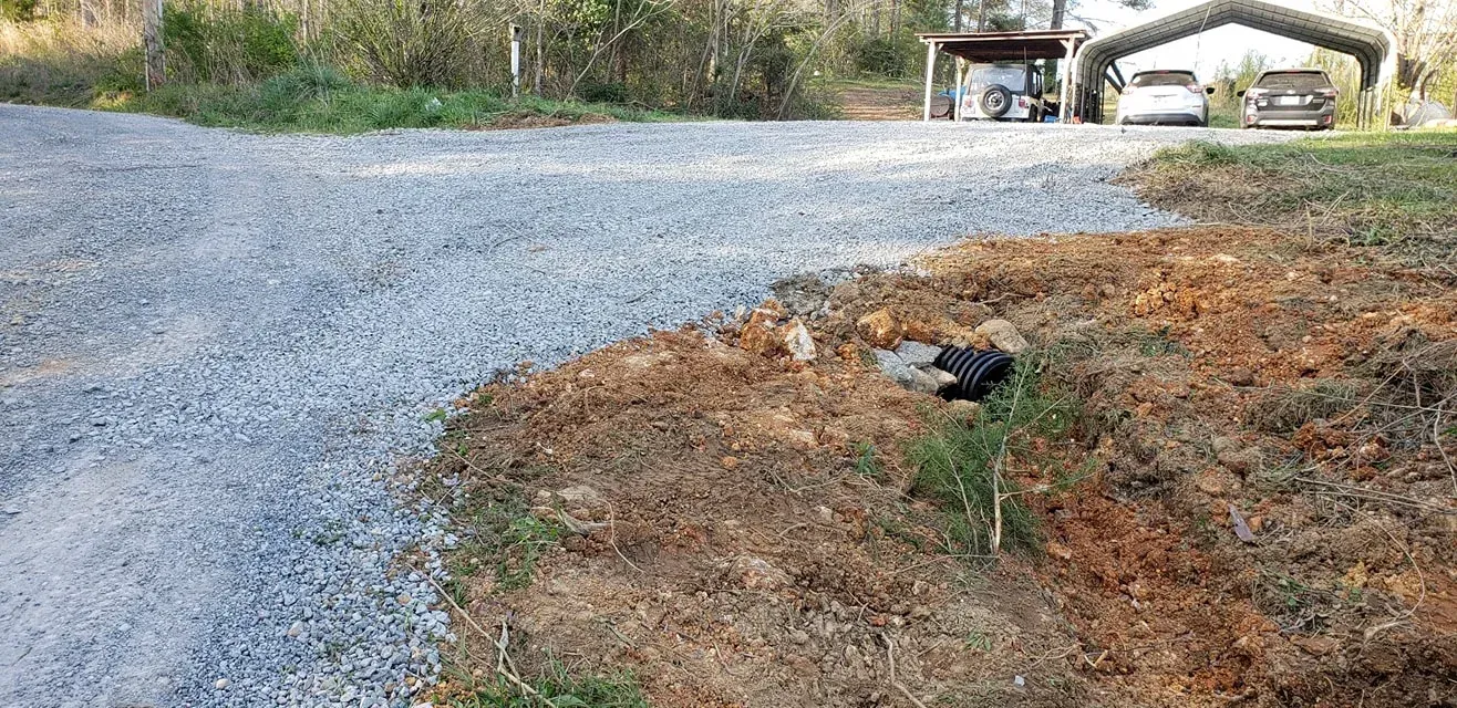Gravel driveway leads to a carport and home. Dirt and grass border the driveway. Black pipe in the grass.