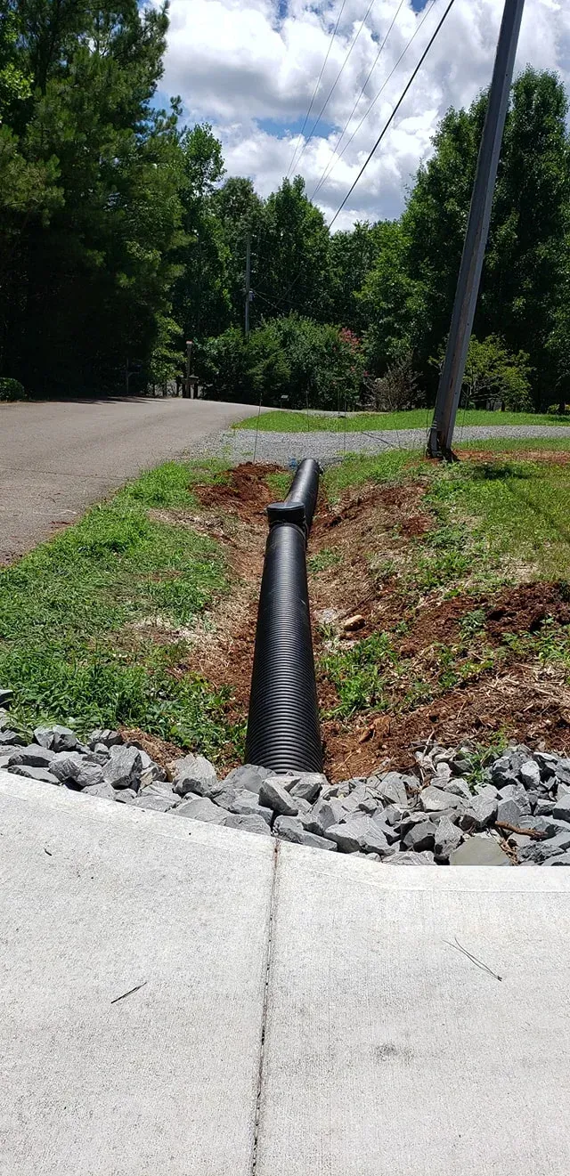 Black corrugated pipe in a ditch beside a concrete sidewalk and dirt, leading to a grassy area and road.