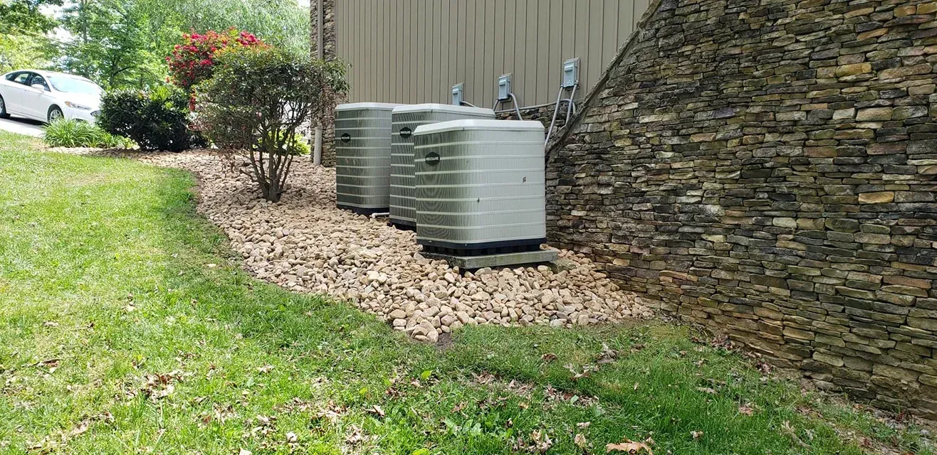Two air conditioning units beside a rock wall and tan siding, with a bed of rocks and grass in the foreground.