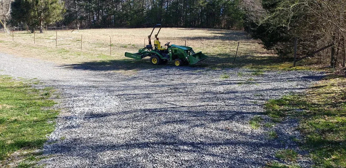 Green tractor on a gravel driveway in front of a field and trees.