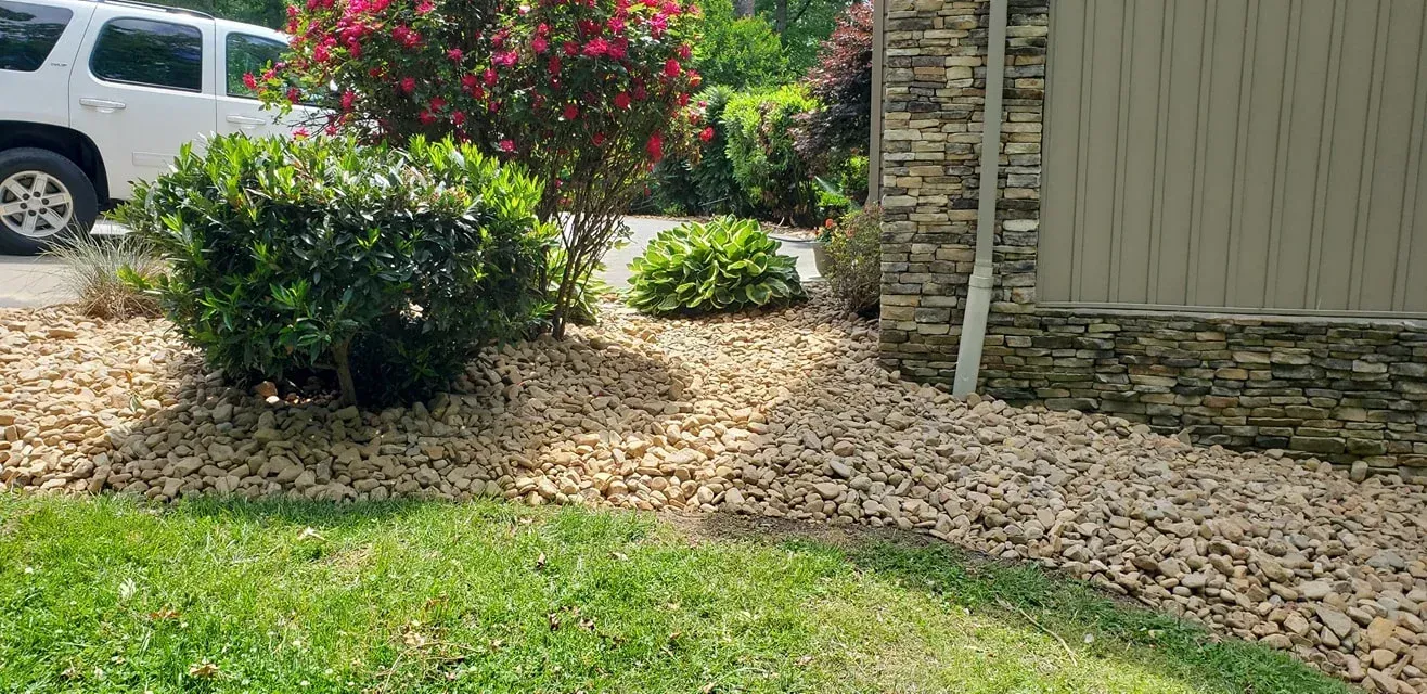 Landscaped front yard with shrubs, rocks, and green grass. A white car and house exterior are visible.