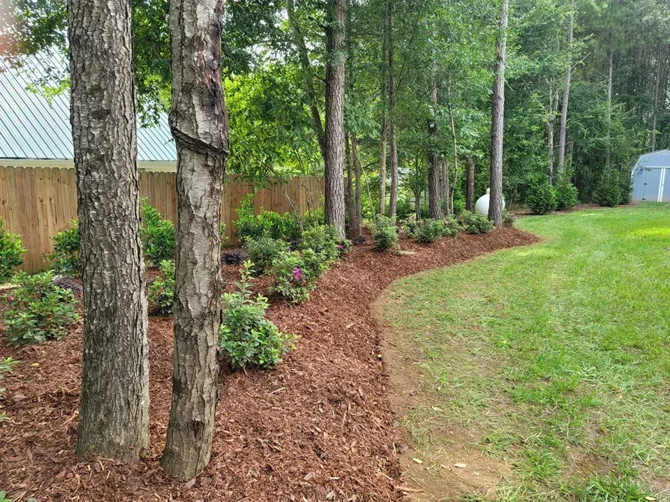 Mulched garden bed borders a green lawn, trees, and wooden fence.