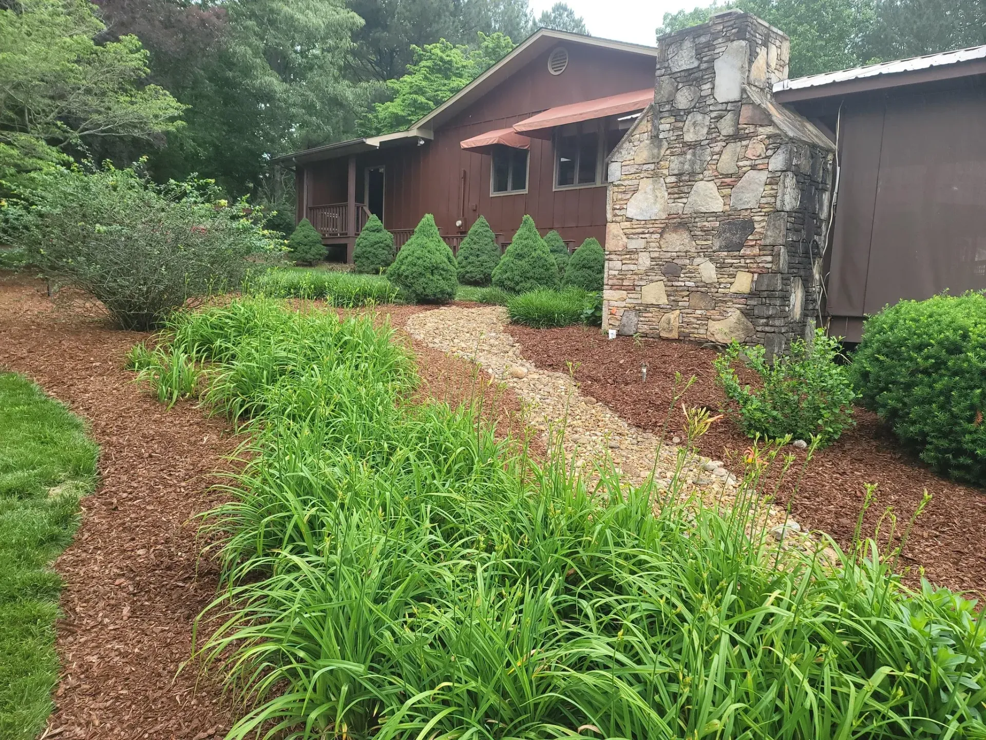 Brown house with stone chimney and garden bed with green plants and mulch.