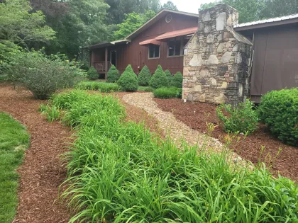 Brown house with stone chimney and garden bed with green plants and mulch.