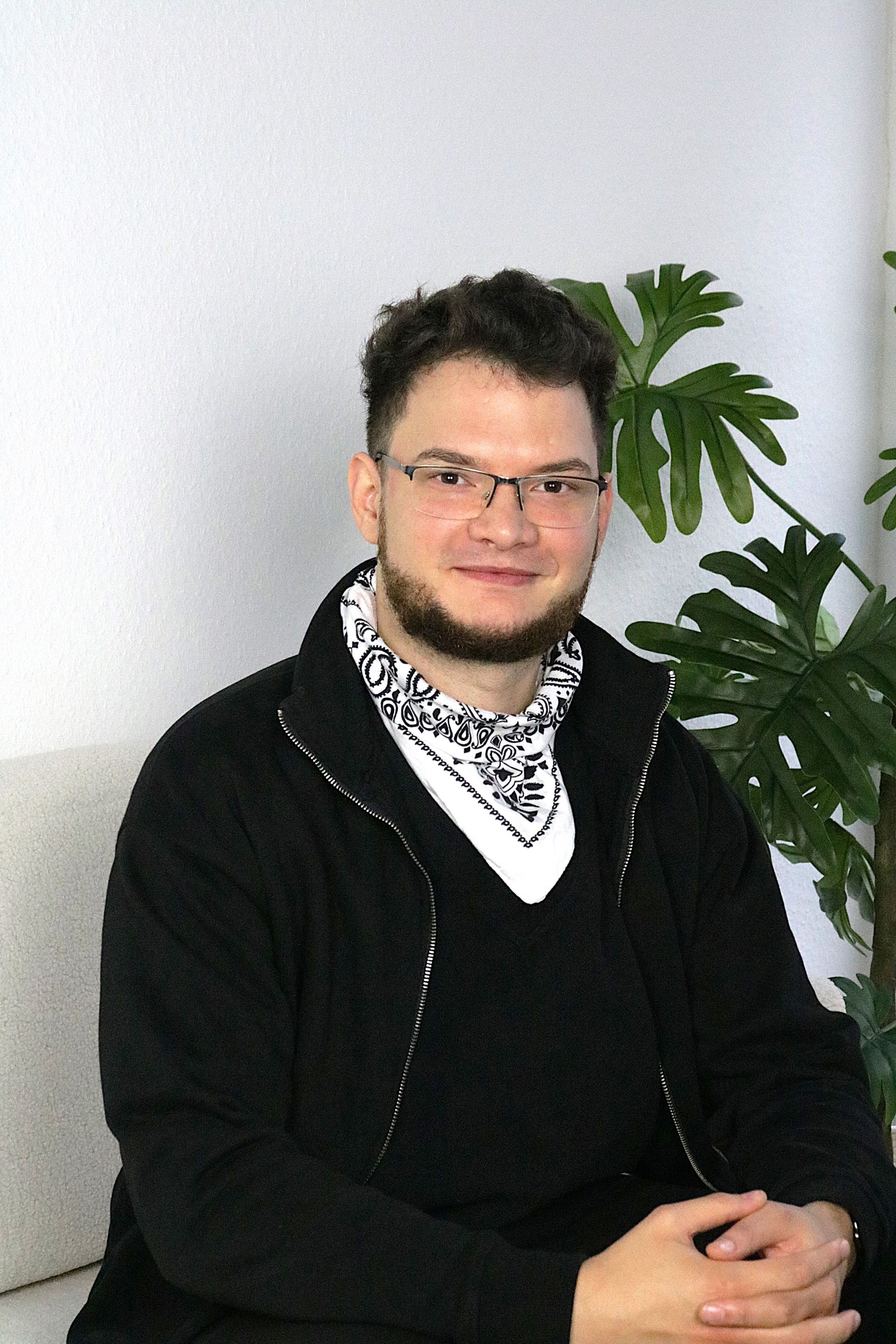 Eine Person in schwarzer Jacke und gemustertem Schal sitzt drinnen neben einer belaubten Pflanze.