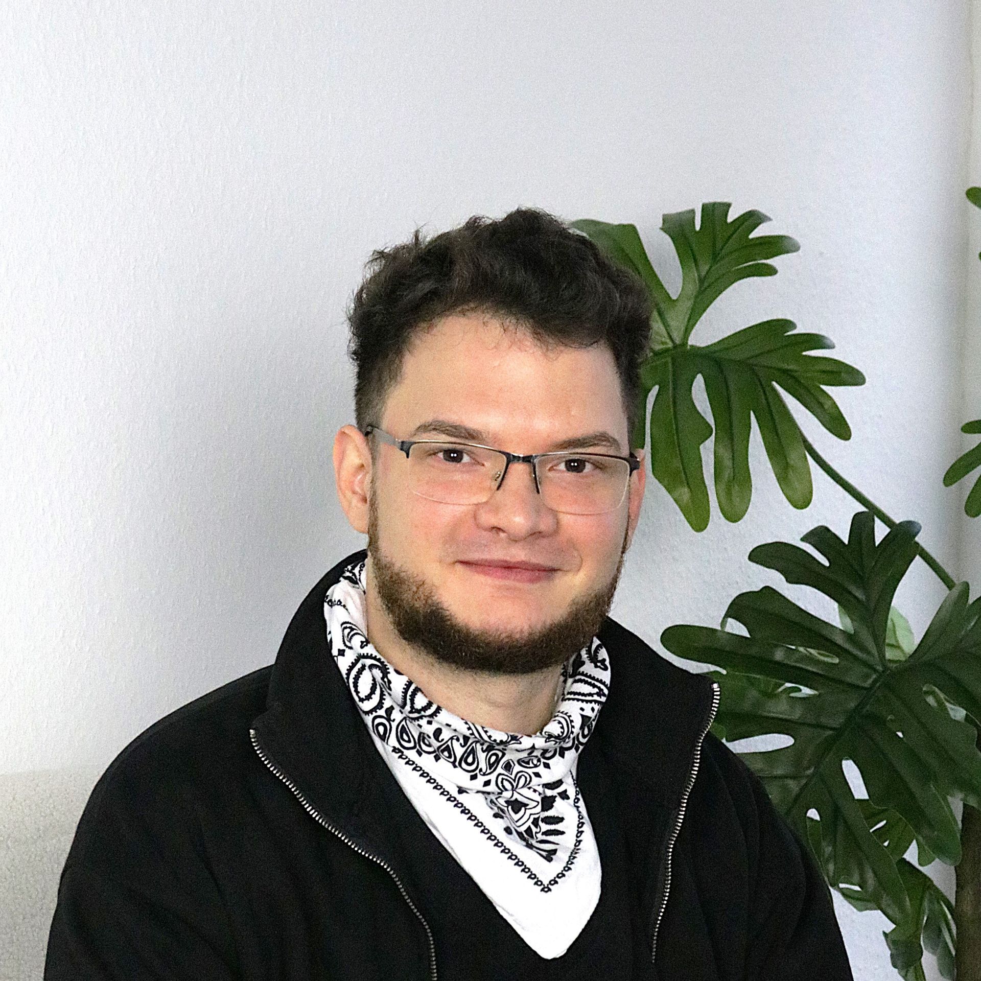 Porträt einer Person mit Brille und schwarzer Jacke, die ein gemustertes Bandana trägt, neben einer belaubten Pflanze.
