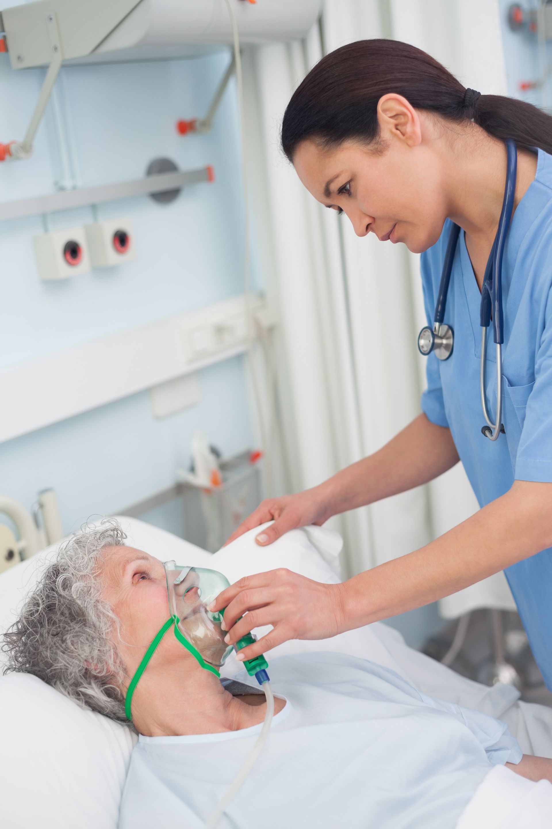 Eine Krankenschwester in blauer OP-Kleidung justiert die Sauerstoffmaske eines Patienten, der in einem Krankenhausbett liegt.