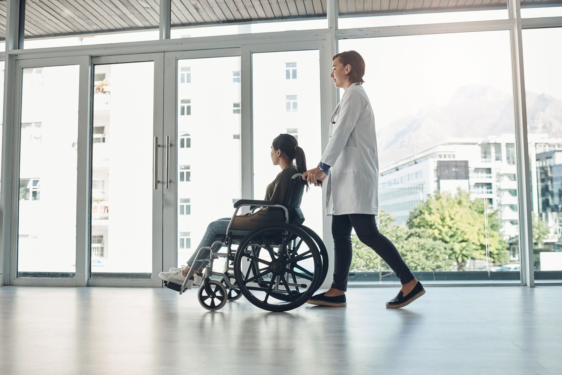 Eine Pflegekraft schiebt eine Person im Rollstuhl durch einen hellen, leeren Flur.