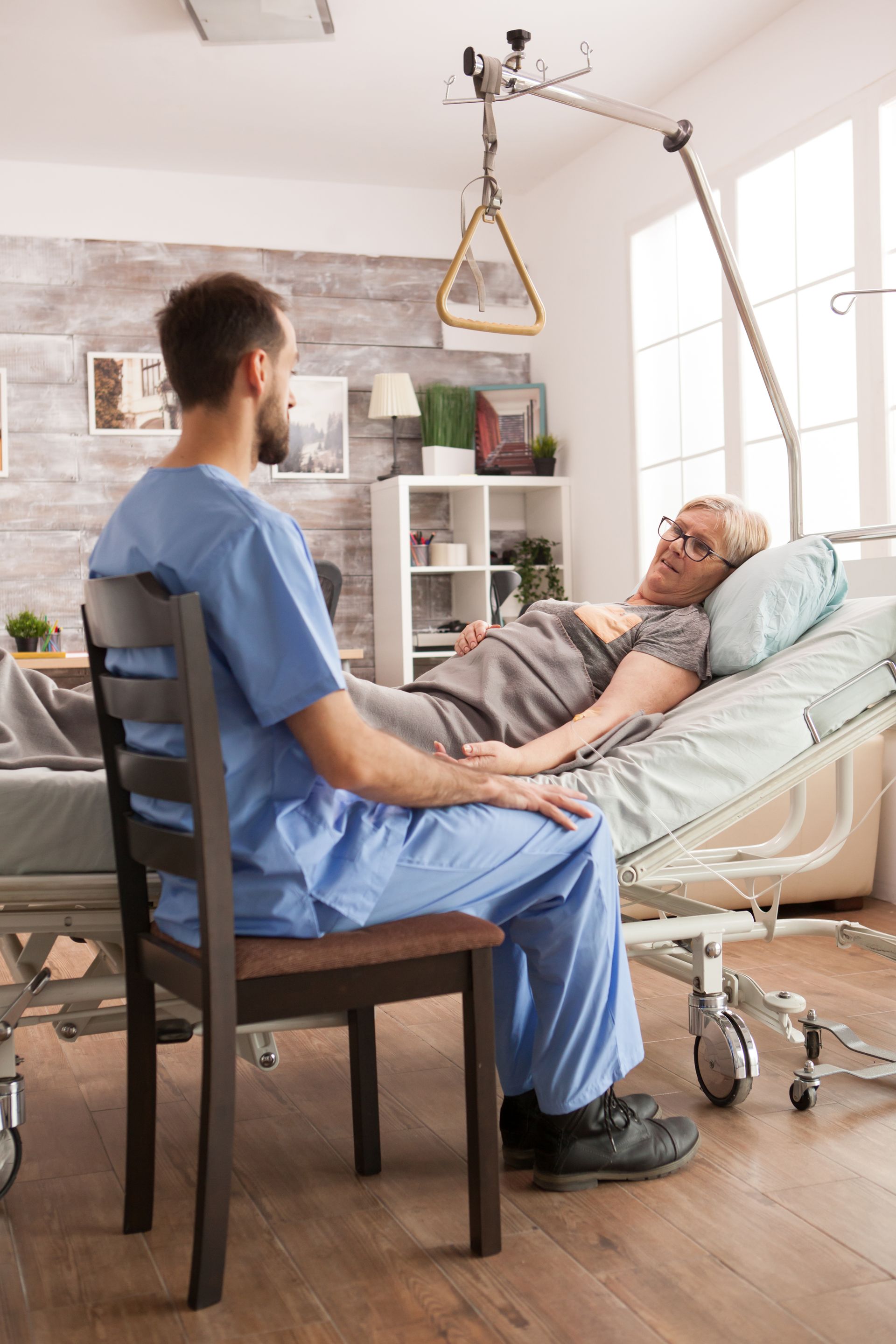 Eine Pfleger in OP-Kleidung sitzt neben einem Patienten in einem Krankenhausbett in einem häuslichen Pflegezimmer.