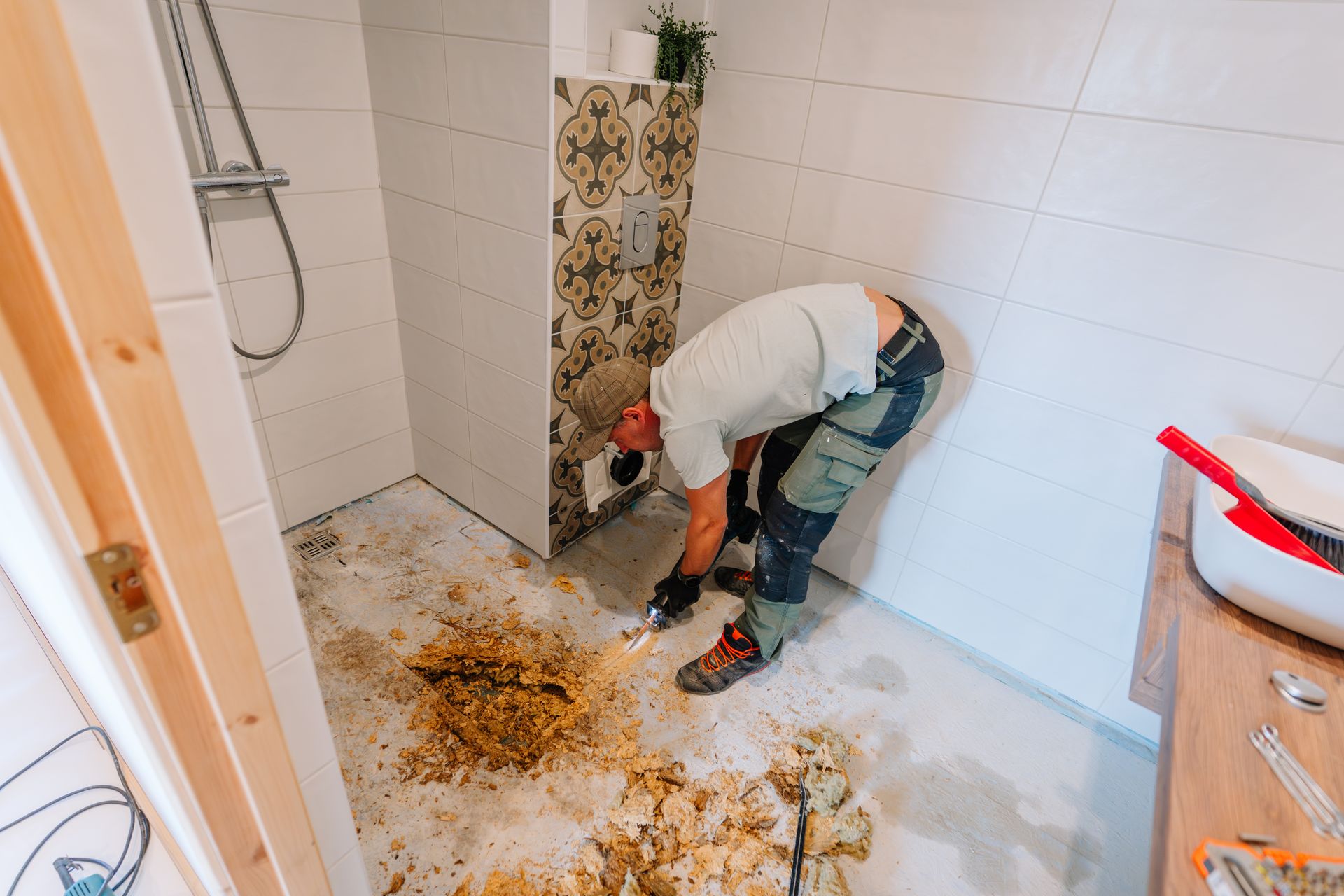 Man working in a bathroom with torn-up flooring, near a shower, white and patterned tile, and wooden trim.