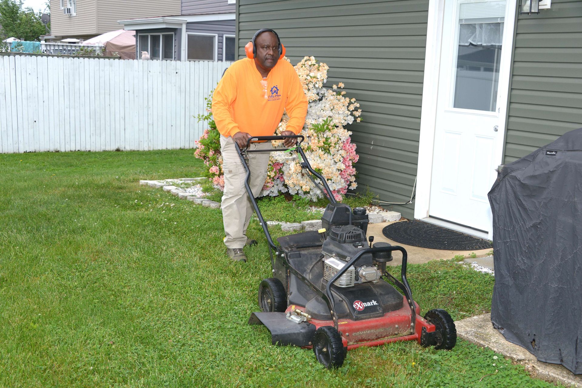A person in an orange hoodie using a push mower in a grassy yard beside a house.