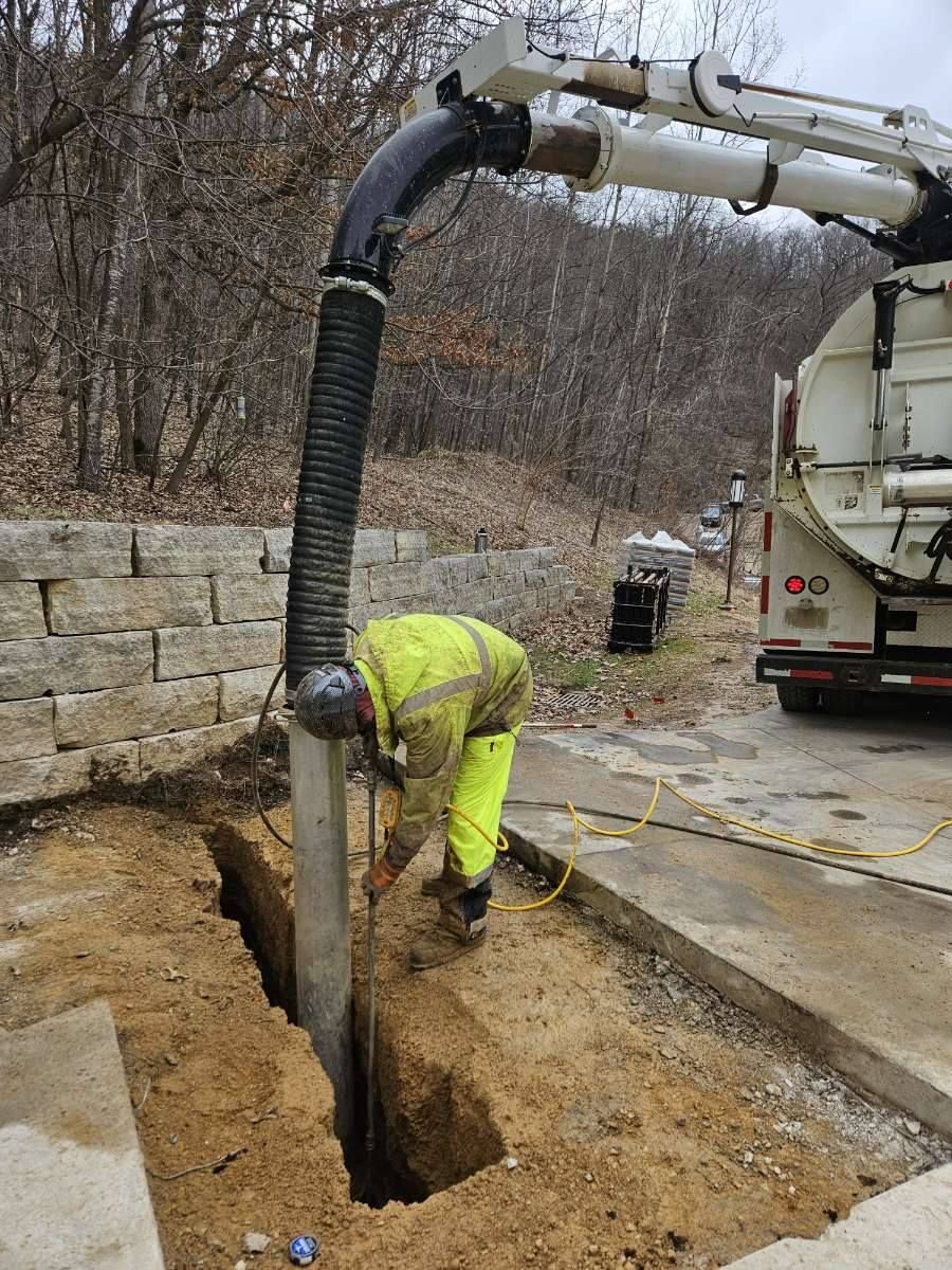 A worker in a high-visibility yellow jacket uses a vacuum excavator truck hose to dig a trench in dirt near a wall.