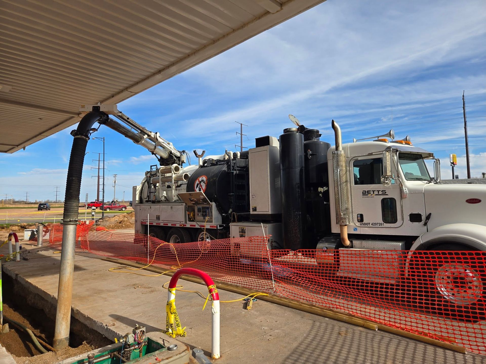 A white vacuum excavation truck operates at a construction site, with its suction hose extended into an underground area.