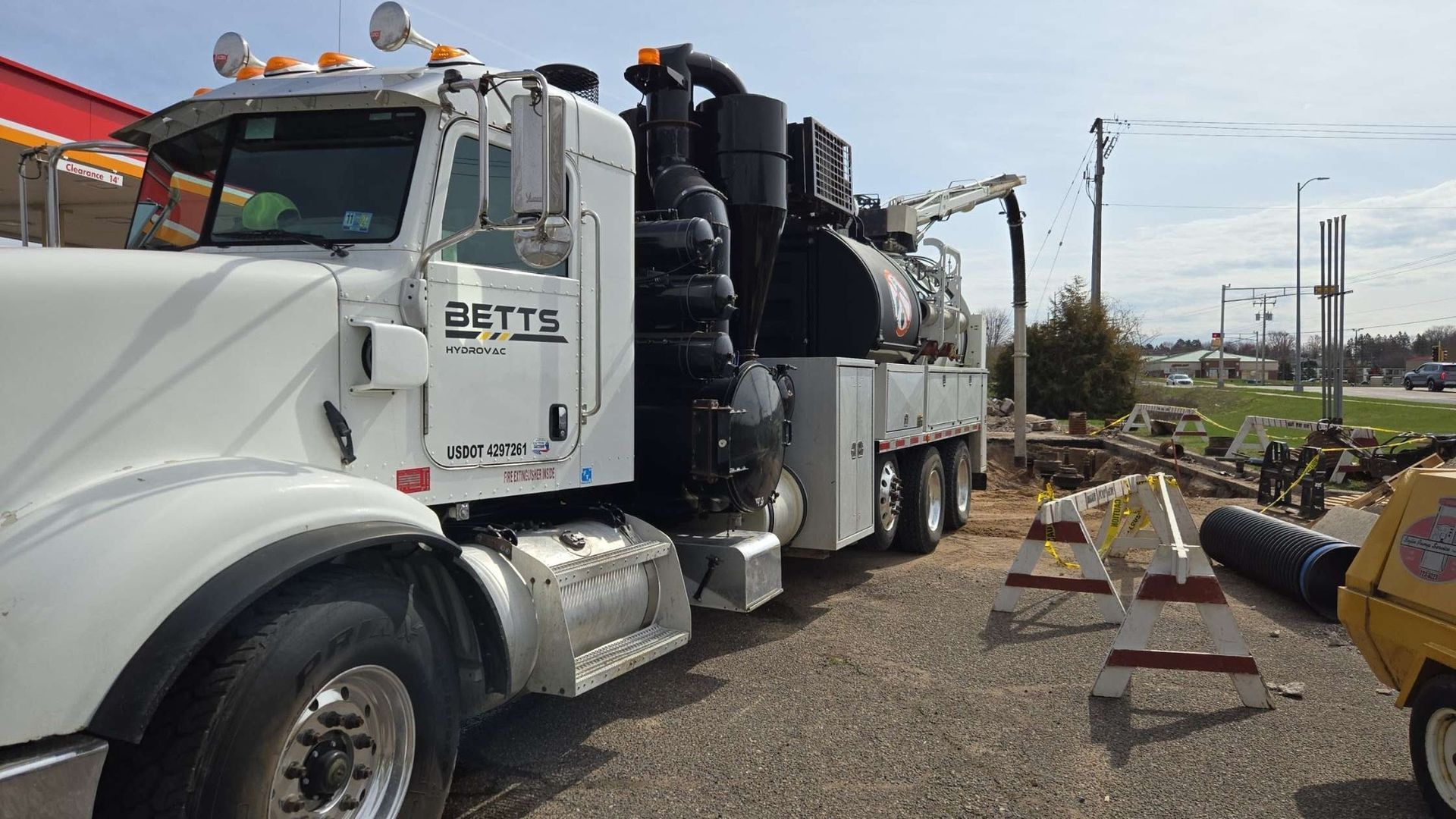 A white vacuum excavation truck parked at a construction site with equipment visible nearby under a bright sky.