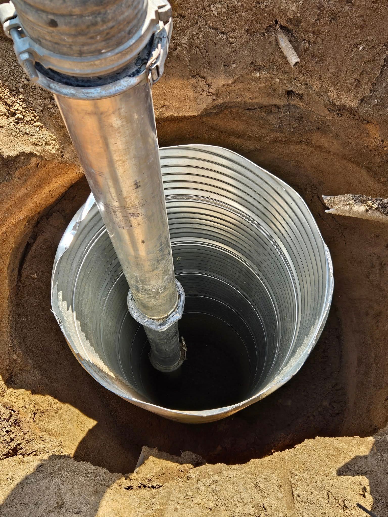 A metal pipe extends vertically into a corrugated metal casing set in a deep, sandy, circular hole in the ground.