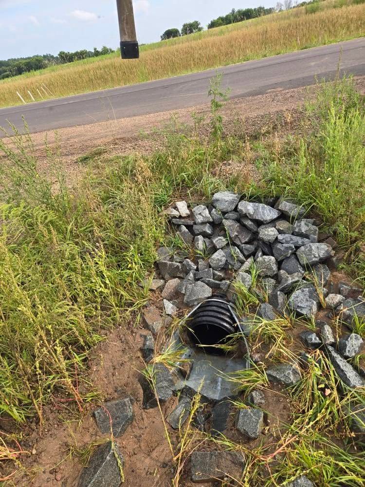 A black corrugated drainage pipe set in a bed of gray rocks next to a road and a grassy field.
