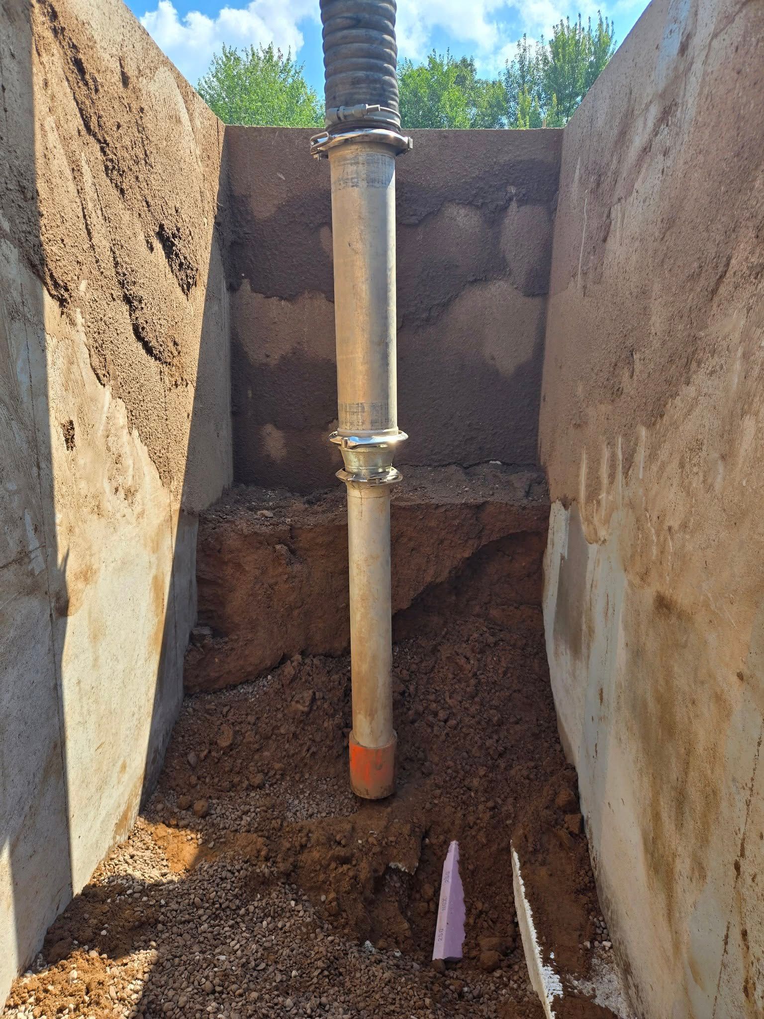 A vertical pipe stands in a dirt-filled construction trench between two rough concrete walls under a bright blue sky.