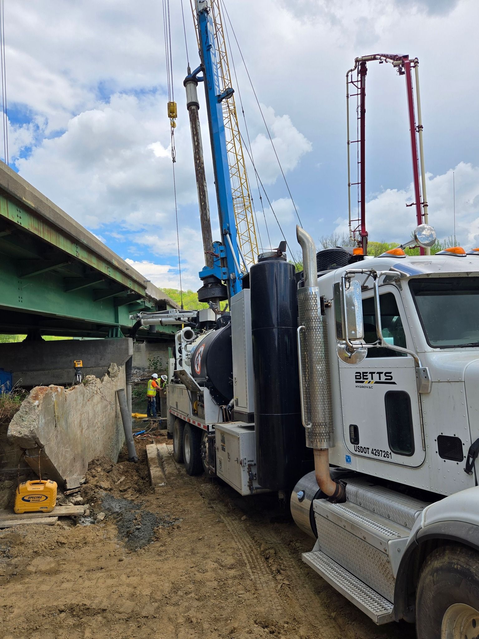 A drilling rig truck parked on a dirt construction site near a bridge overpass under a cloudy sky.
