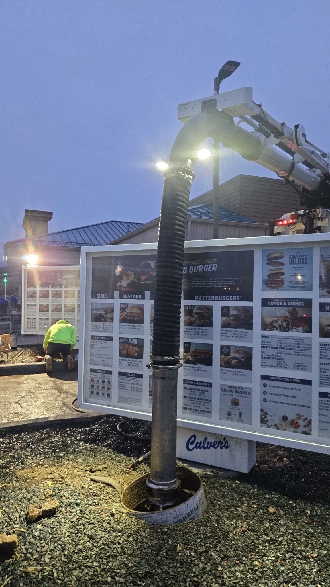 A large industrial vacuum hose is positioned over a fast-food menu board at dusk, with a worker visible nearby.