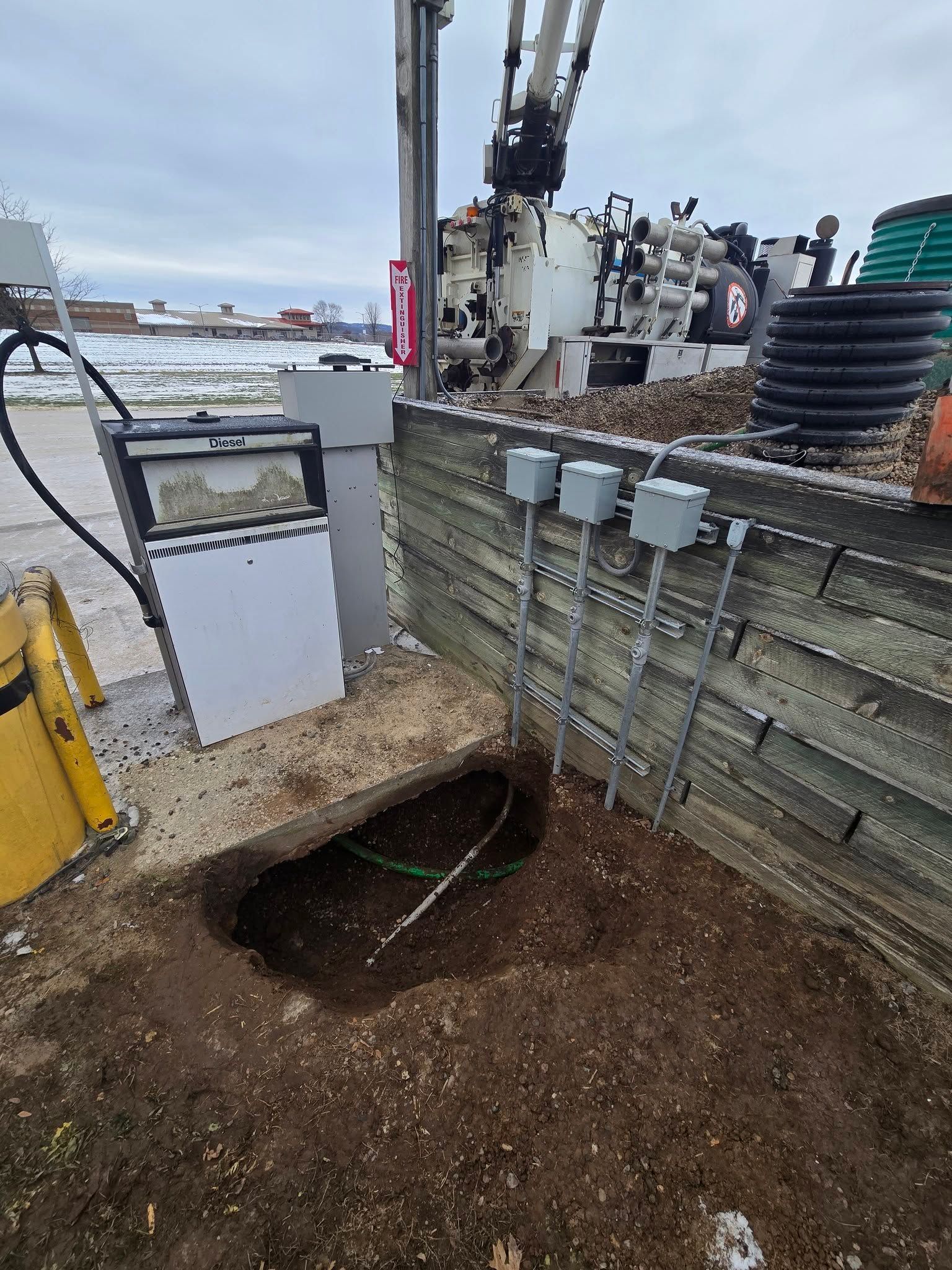 A construction site featuring an exposed underground trench, three electrical boxes on a timber wall, and a crane nearby.