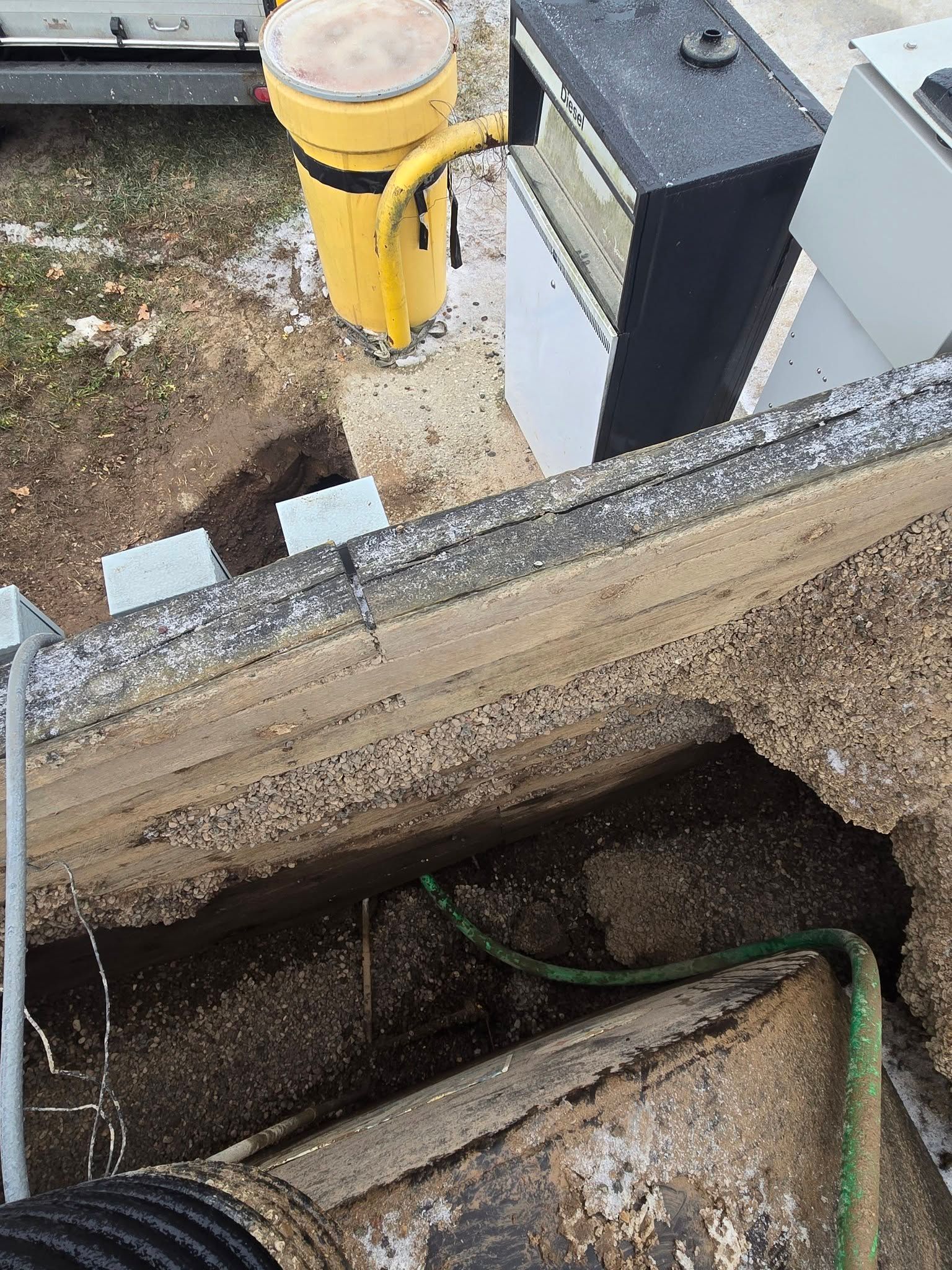 A construction site shows a deep trench next to a concrete ledge, a yellow pipe, and a black electrical box.