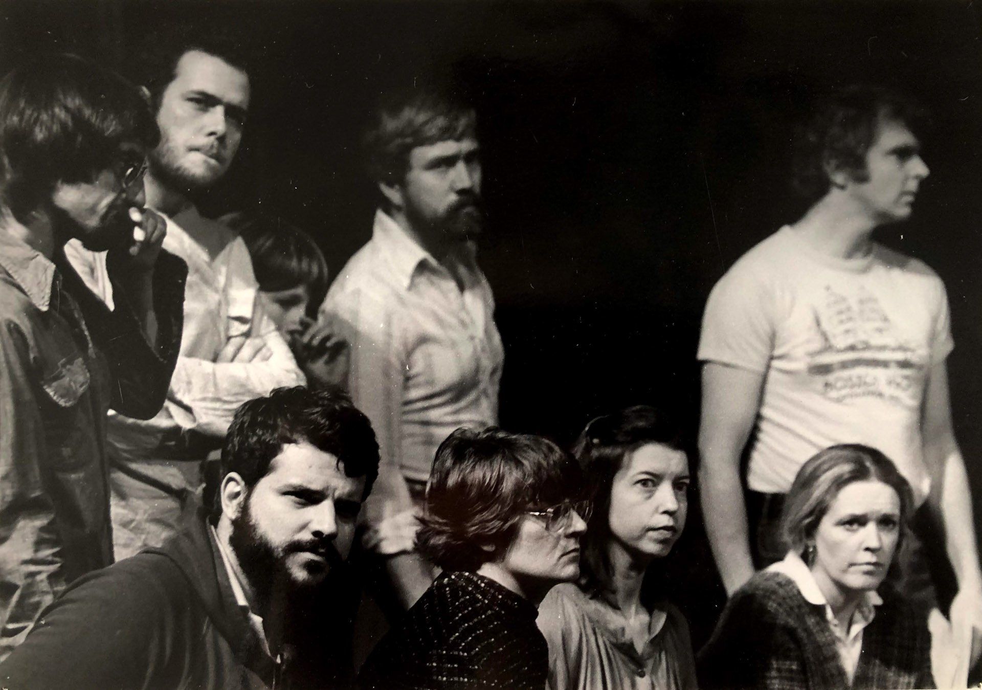 Group of people, mostly men, looking to the side. Some have beards, wearing casual clothing, likely outdoors.