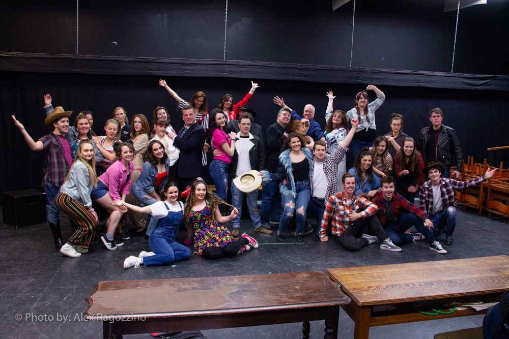 Group of people posing on a stage, some wearing costumes. Wooden tables in the foreground.