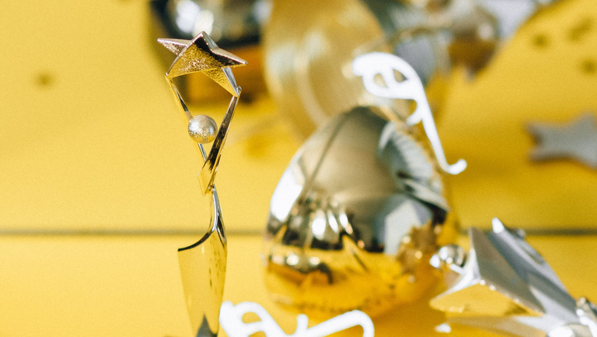 Gold trophy with star and pearl, surrounded by gold and silver decorations on yellow surface.