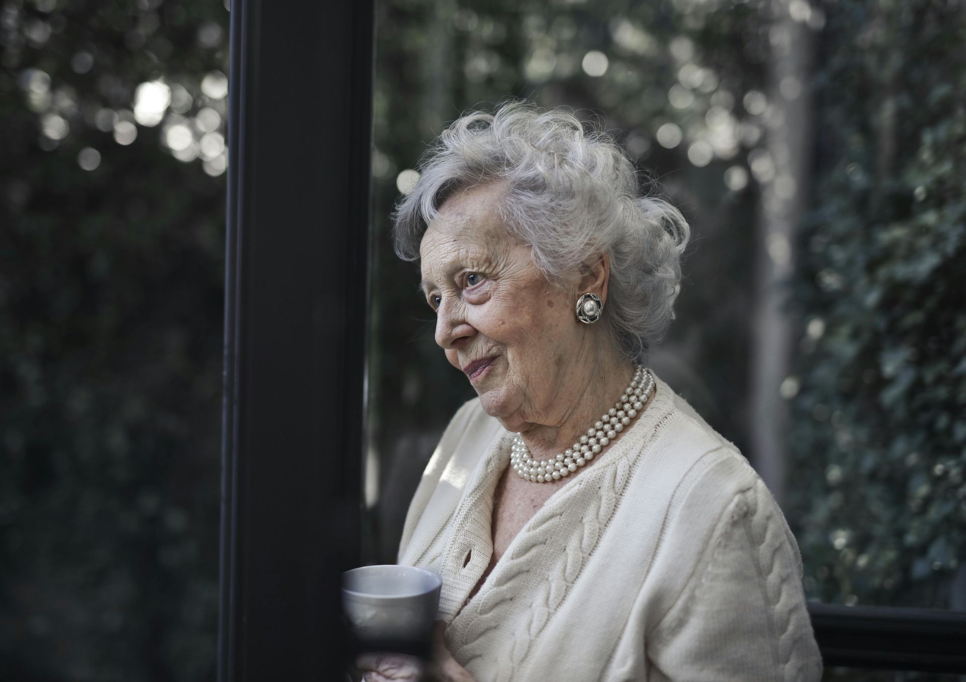 An elderly woman is standing on a porch holding a cup of coffee.