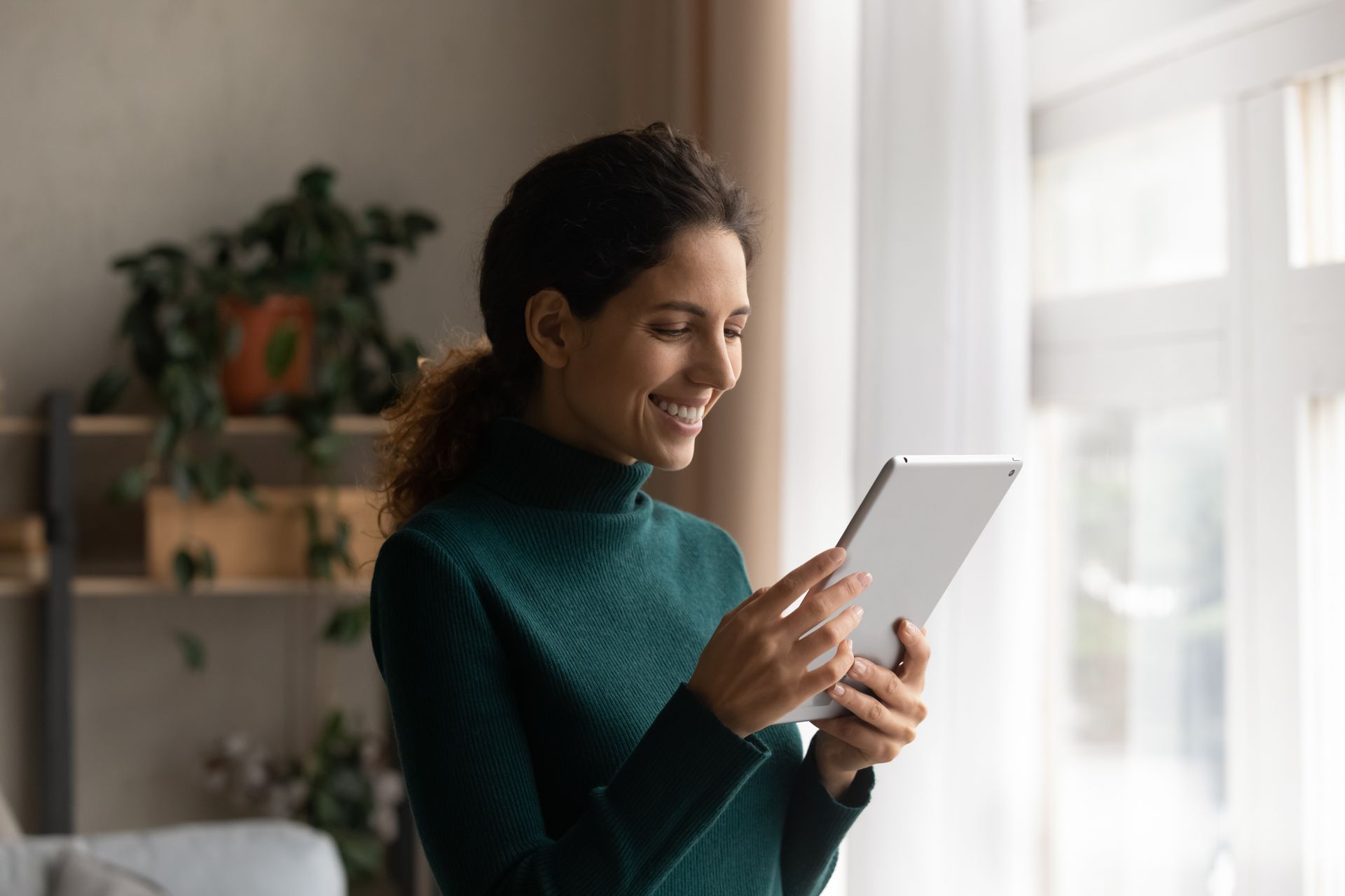 A woman is smiling while using a tablet computer in front of a window.