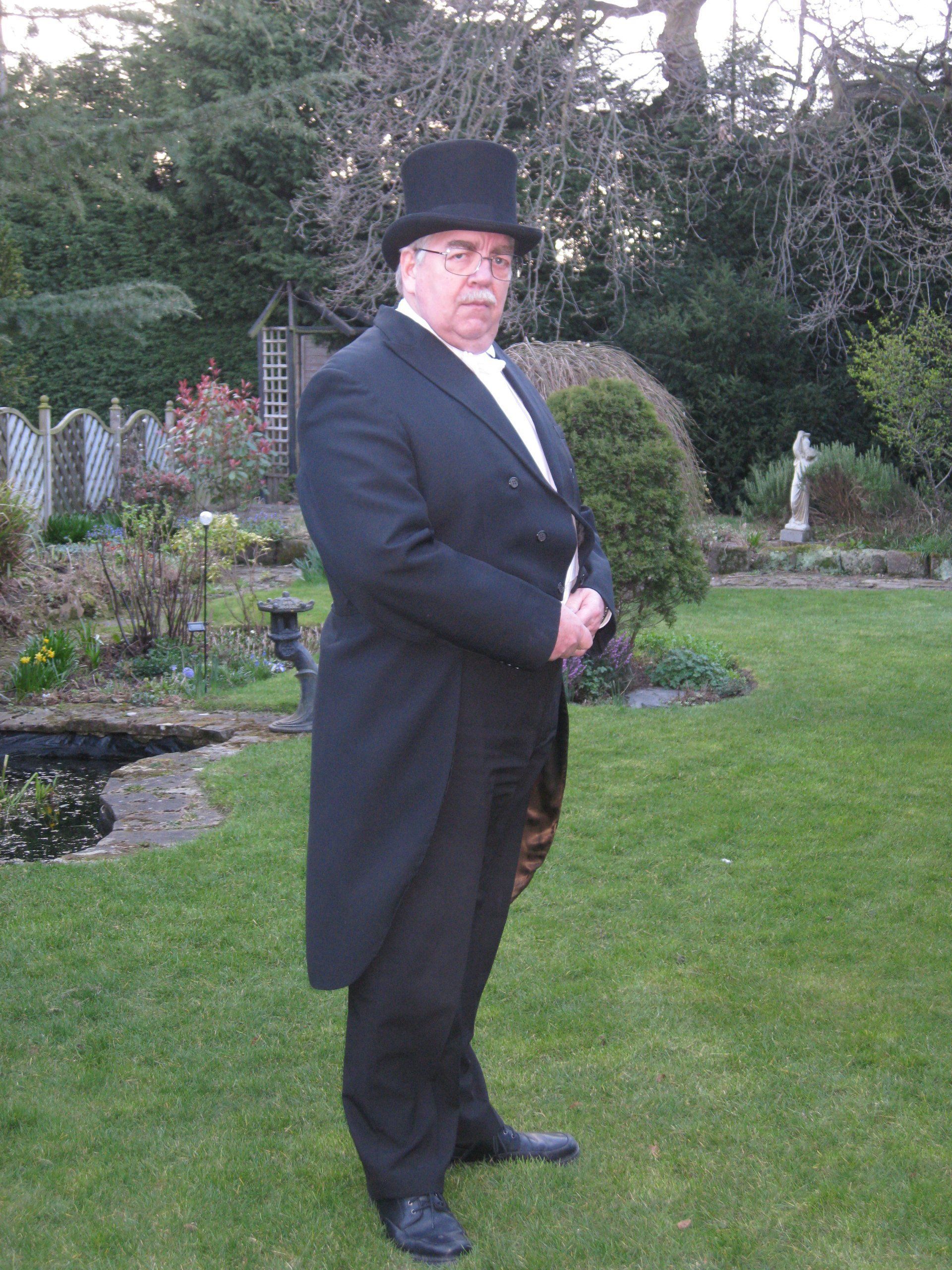 A man in a black suit and top hat is standing in a garden.