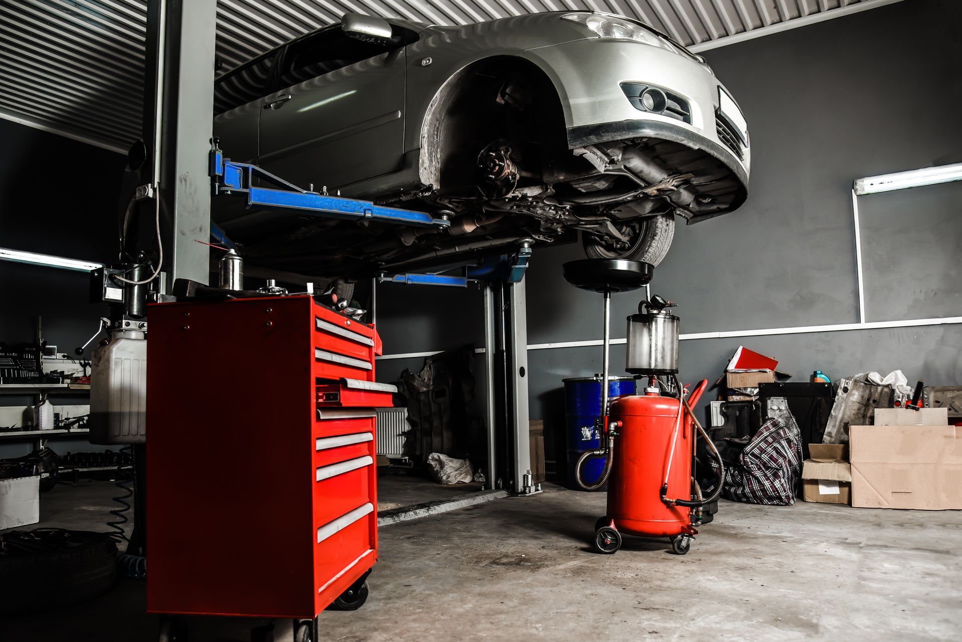 Car on a lift in a garage, undergoing maintenance. Red toolbox and oil drain nearby.