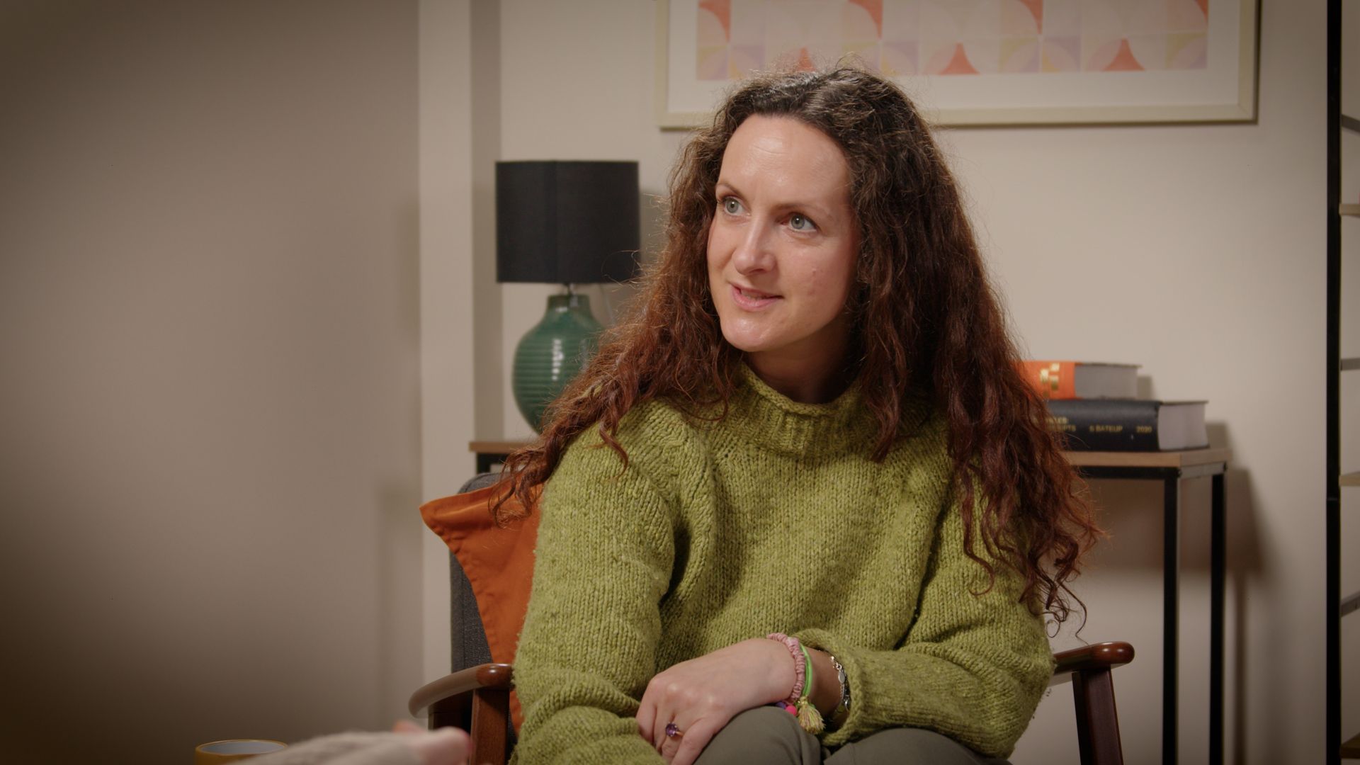 Woman in a green sweater seated indoors, looking to the side with a slight smile.