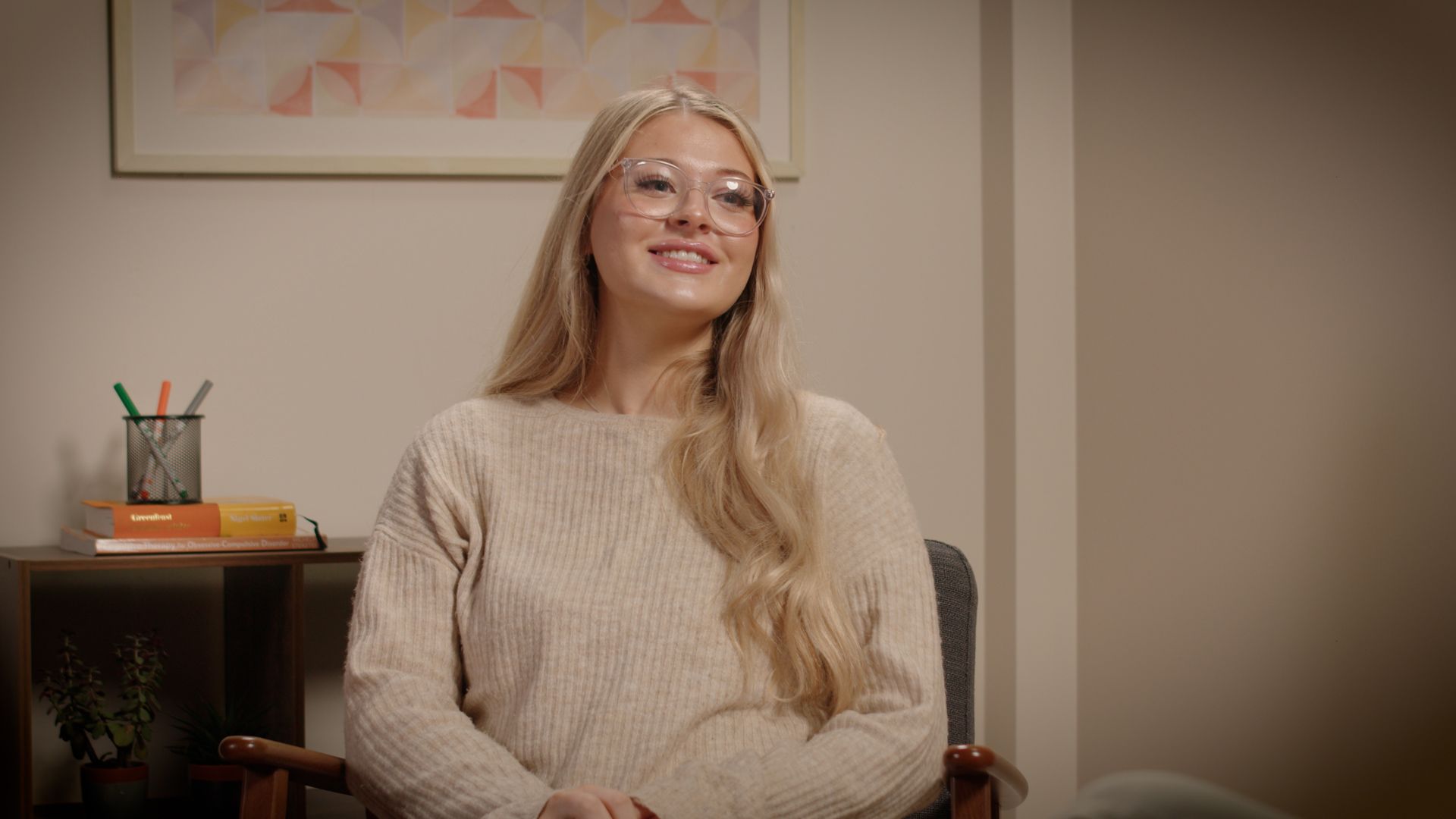 Woman smiling in glasses, seated indoors beside a shelf with pencils and a framed wall print