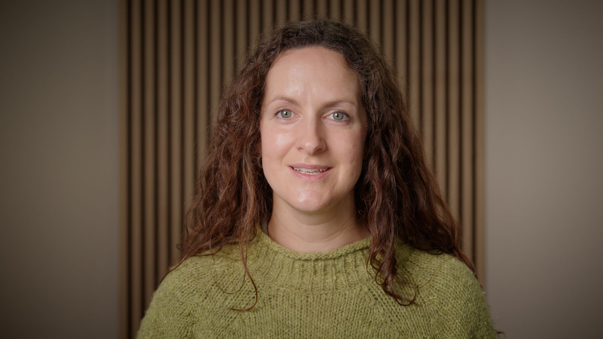 Portrait of a smiling person with curly hair in a green sweater against a striped backdrop