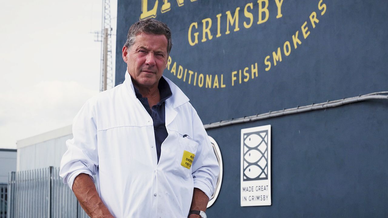 Man in white coat standing by a Grimsby traditional fish smokers sign