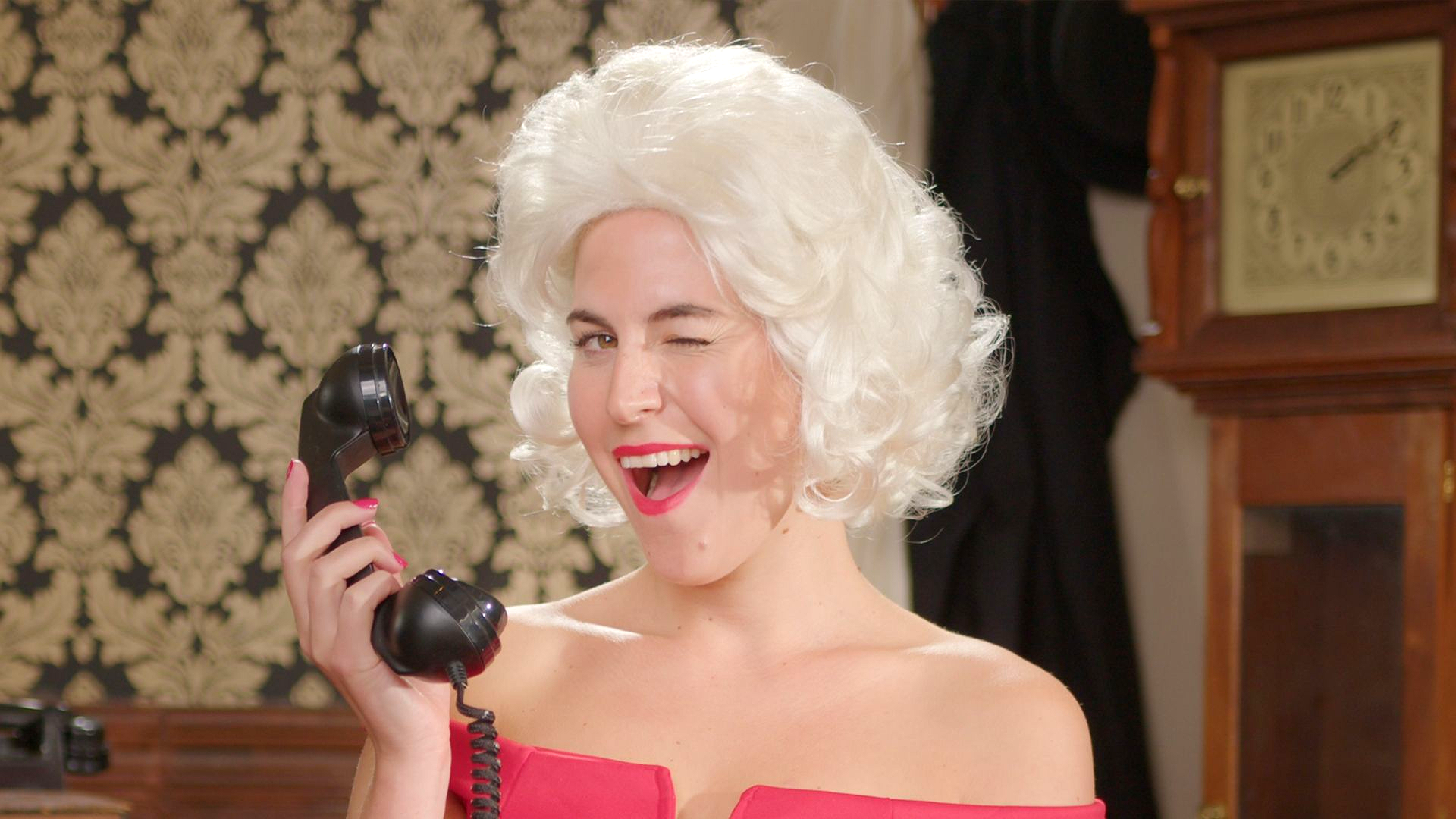 Person in a white wig and red dress laughing while holding a black vintage telephone indoors.