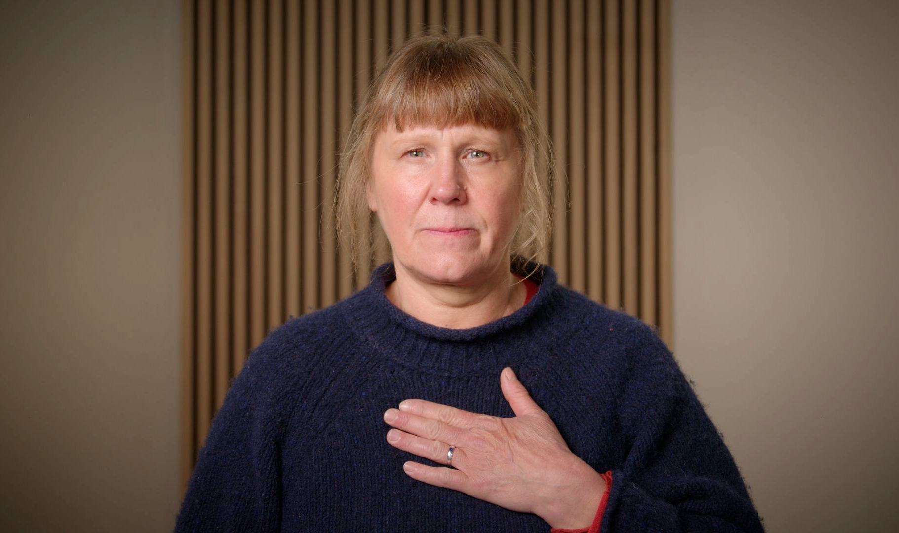 Person in a navy sweater with a hand on their chest, standing against a striped beige background.