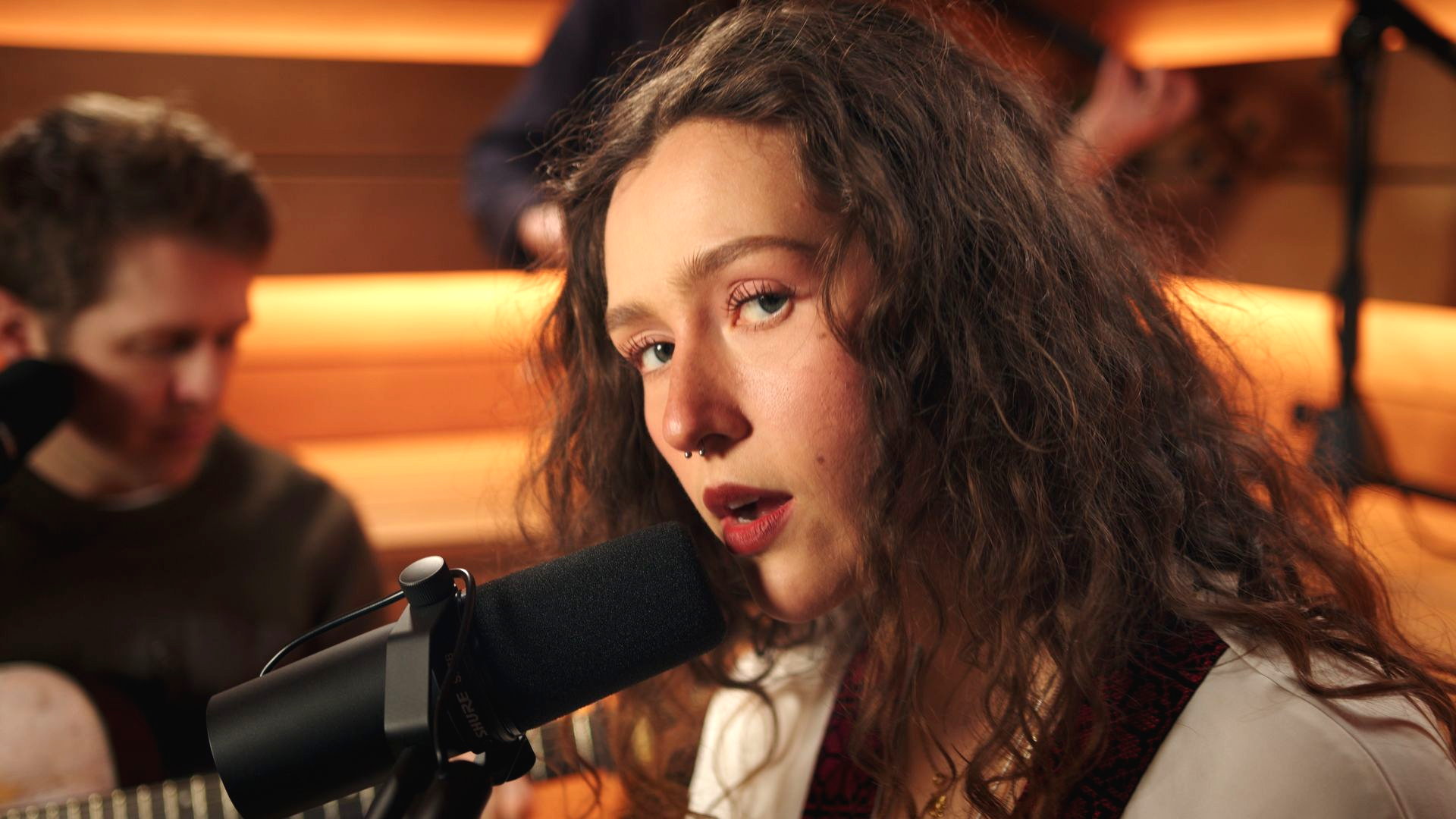 Woman singing into a microphone in a warm-lit recording studio, with a blurred guitarist in the background