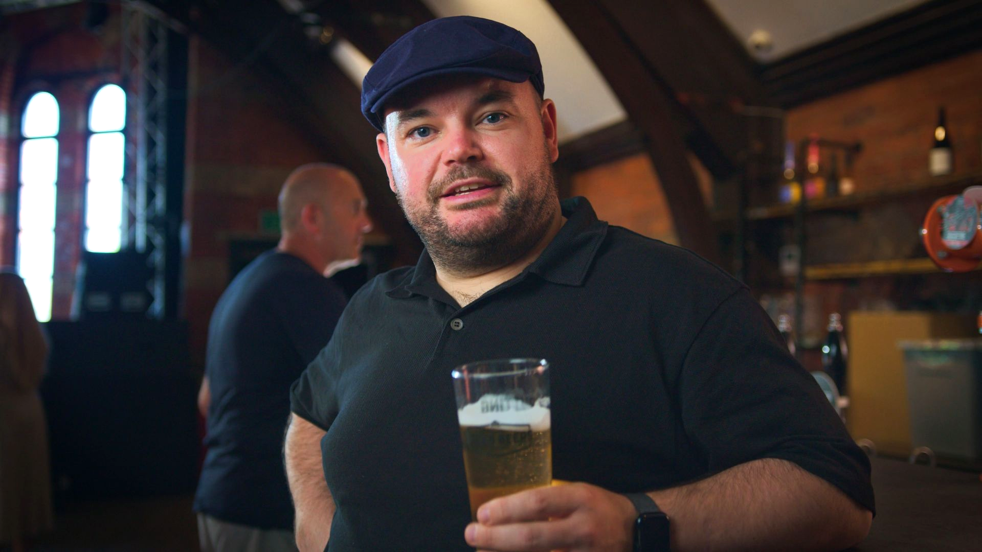 Man in a black shirt and cap holds a pint in a dimly lit bar, looking at the camera.