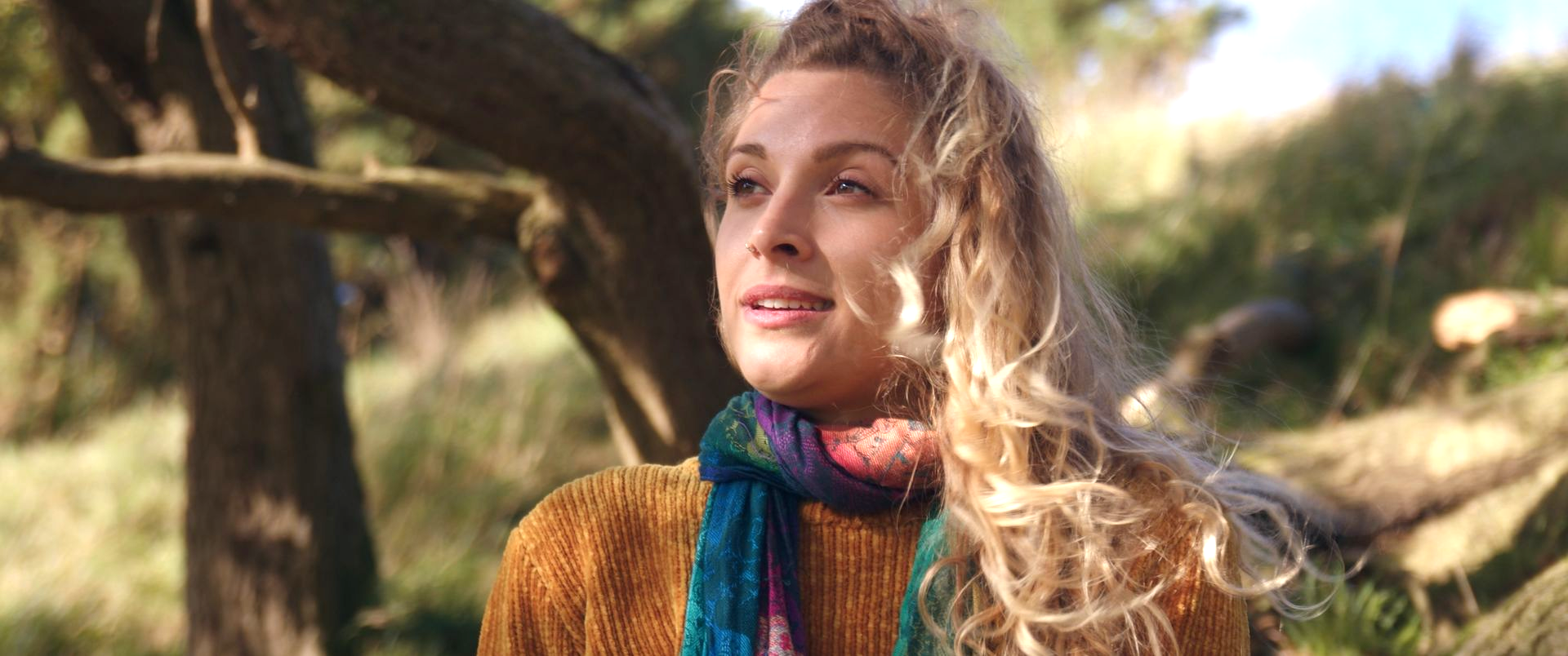 Woman with long blonde hair and a colorful scarf standing outdoors in a sunlit natural setting