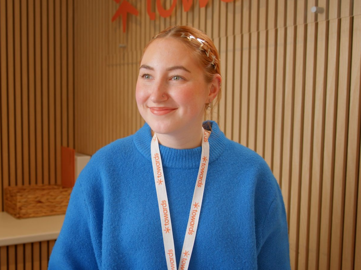 Smiling person in a blue sweater with a lanyard, standing in front of a wooden slat wall.