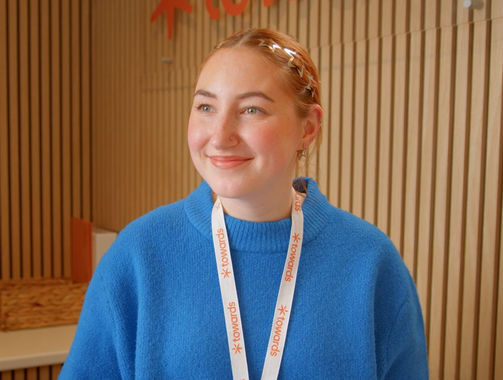 Smiling person in a blue sweater and lanyard standing in a wood-paneled room