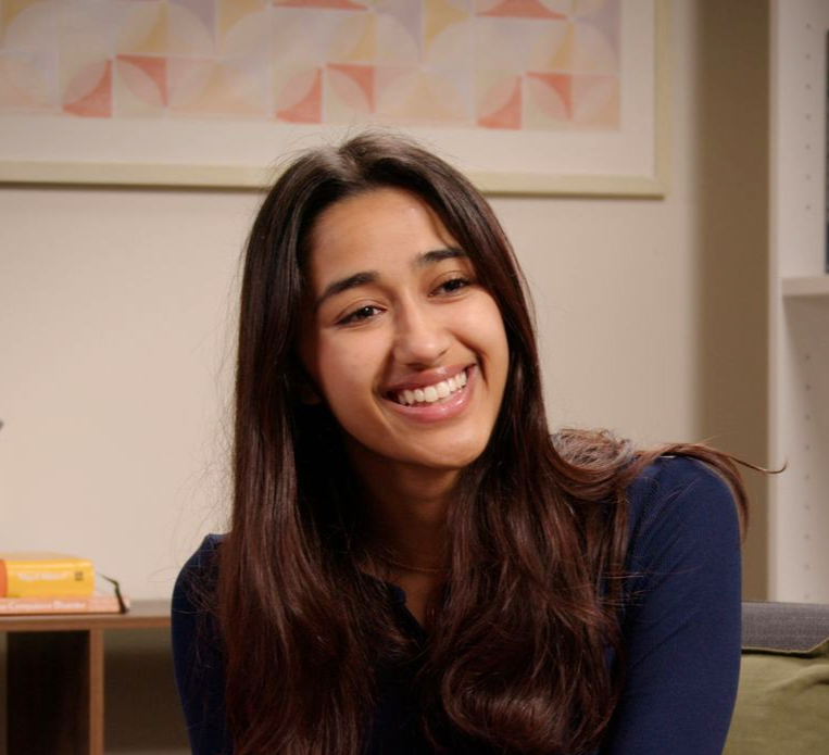 Smiling woman with long dark hair sitting indoors in a room with framed wall art.